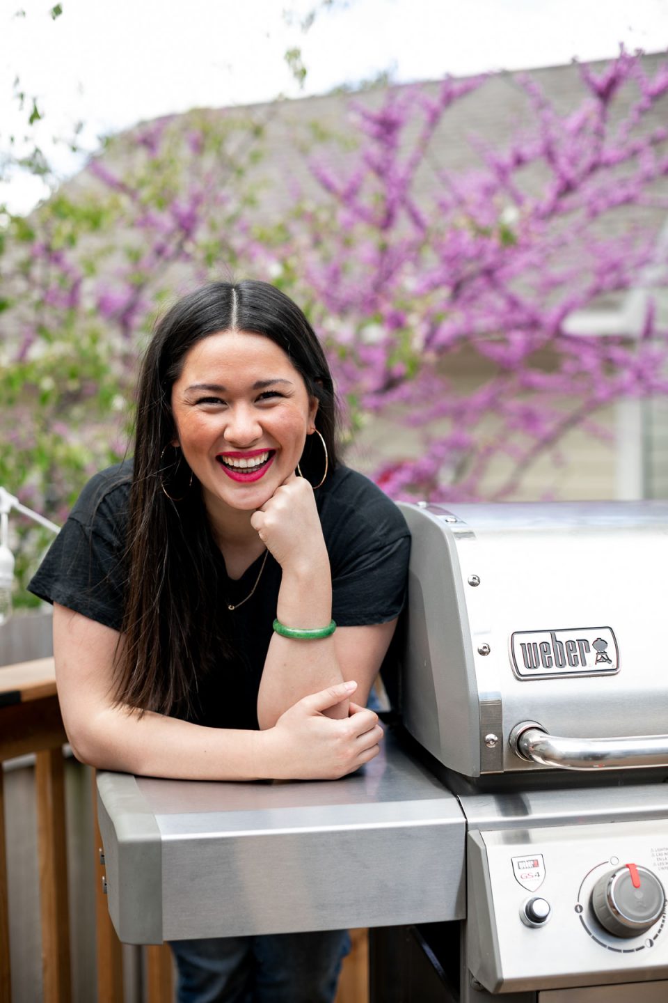 Jess of Plays Well With Butter stands on a deck behind a stainless steel Weber Genesis II propane grill. In the background, a blossoming tree & a house with beige siding.
