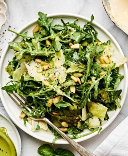 A fresh arugula salad with shaved Parmesan, pine nuts, and a light green dressing is served on a white plate with a fork. The salad is garnished with cracked black pepper.