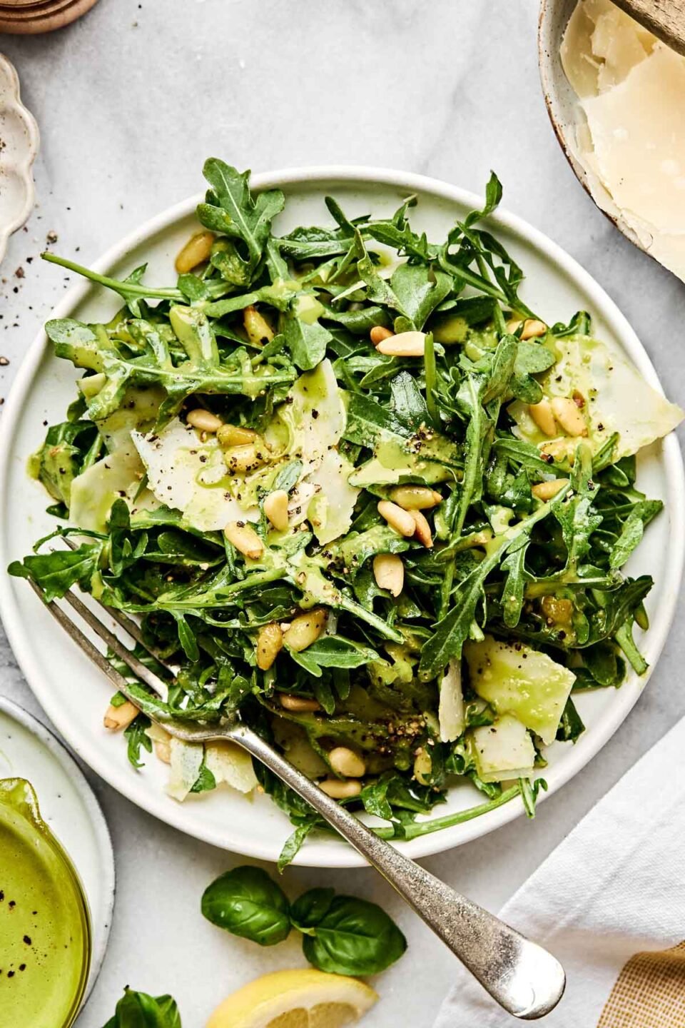 A fresh arugula salad with shaved Parmesan, pine nuts, and a light green dressing is served on a white plate with a fork. The salad is garnished with cracked black pepper.