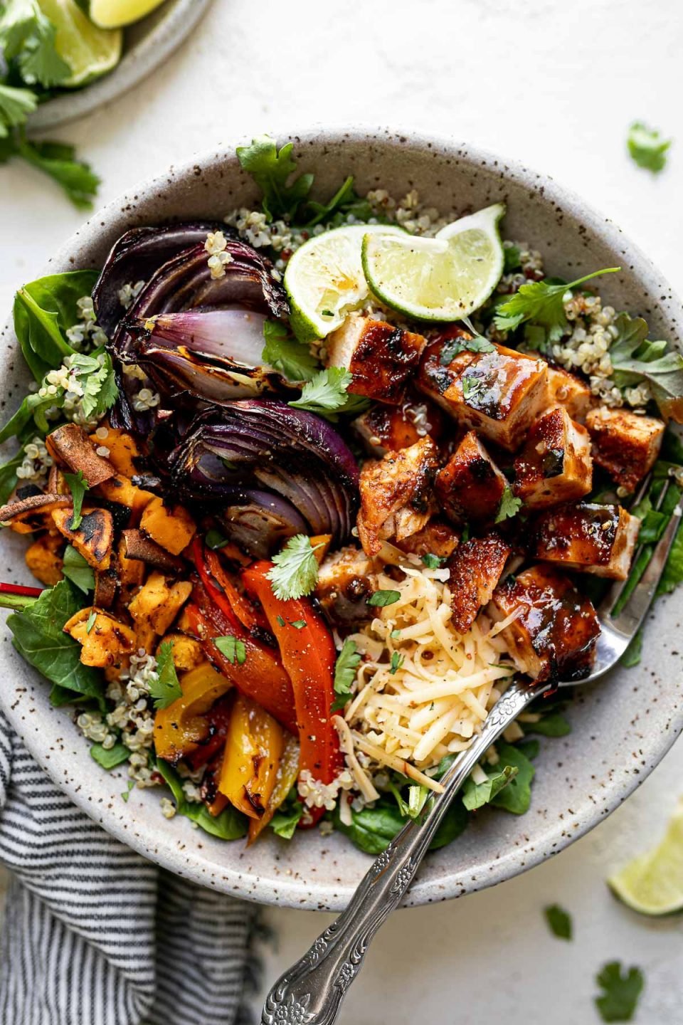 Grilled BBQ chicken bowl with diced BBQ chicken, grilled onions, sweet potatoes and bell peppers sitting atop a bed of greens and quinoa in a large speckled gray bowl. The bowl sits atop a white surface, surrounded by a striped gray linen napkin, cilantro leaves and lime wedges.