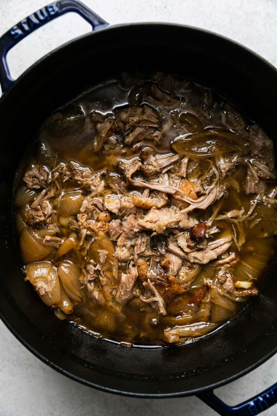 Cooked and shredded pork carnitas in a beer braise fill a dark navy Staub dutch oven. The dutch oven sits atop a creamy white textured surface.