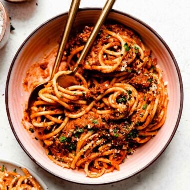An overhead shot of pasta bolognese in a large white bowl atop a white surface. The bowl sits alongside a second bowl of pasta and dishes of salt and red pepper flakes.