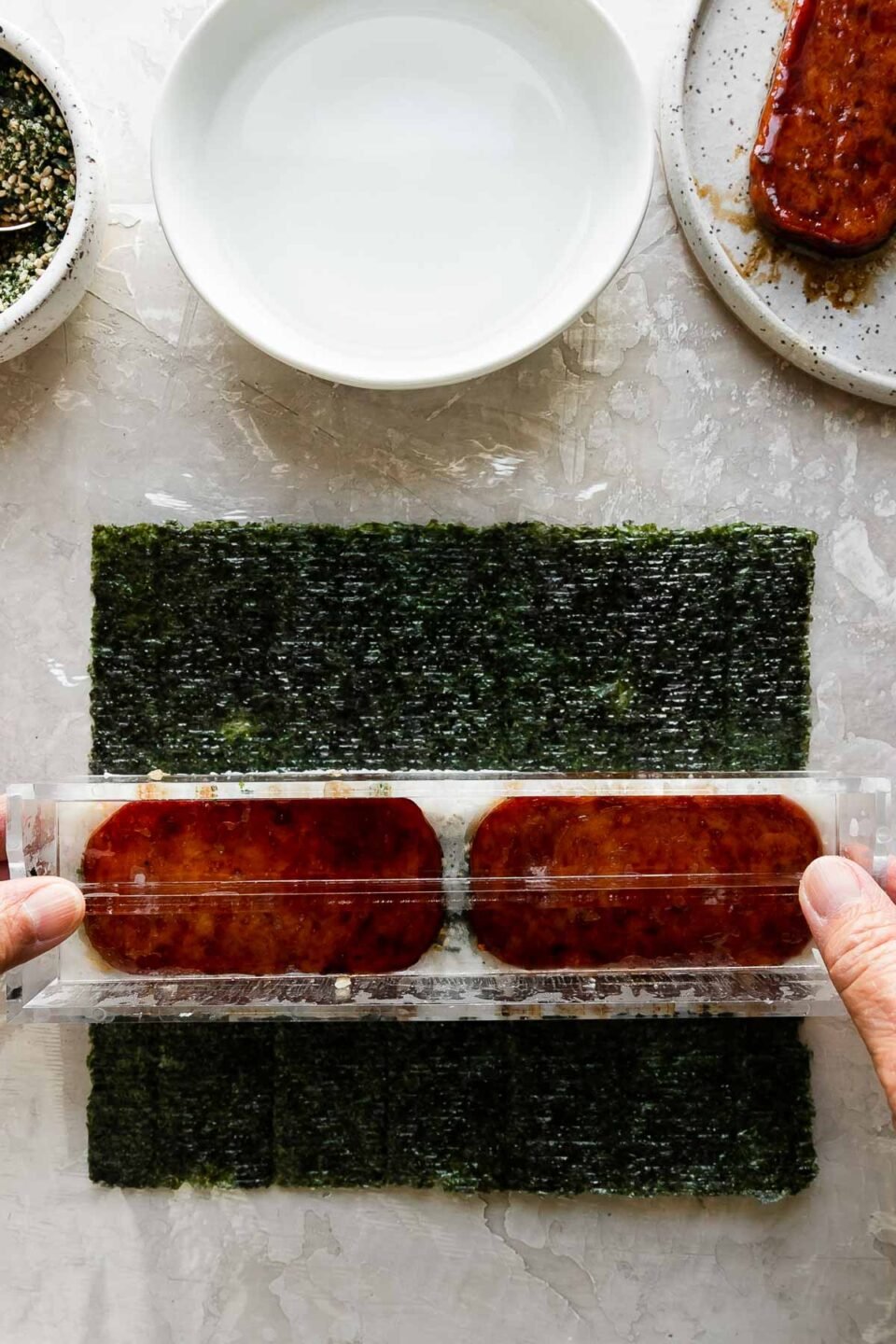 A close up of a woman using her hands to remove the lid of a spam musubi filled musubi mold to create a finished spam musubi. Resting above the musubi mold is a small bowl filled with furikake seasoning with a spoon resting inside, another small white bowl filled with water, and a small speckled ceramic plate with pan-fried teriyaki Spam.