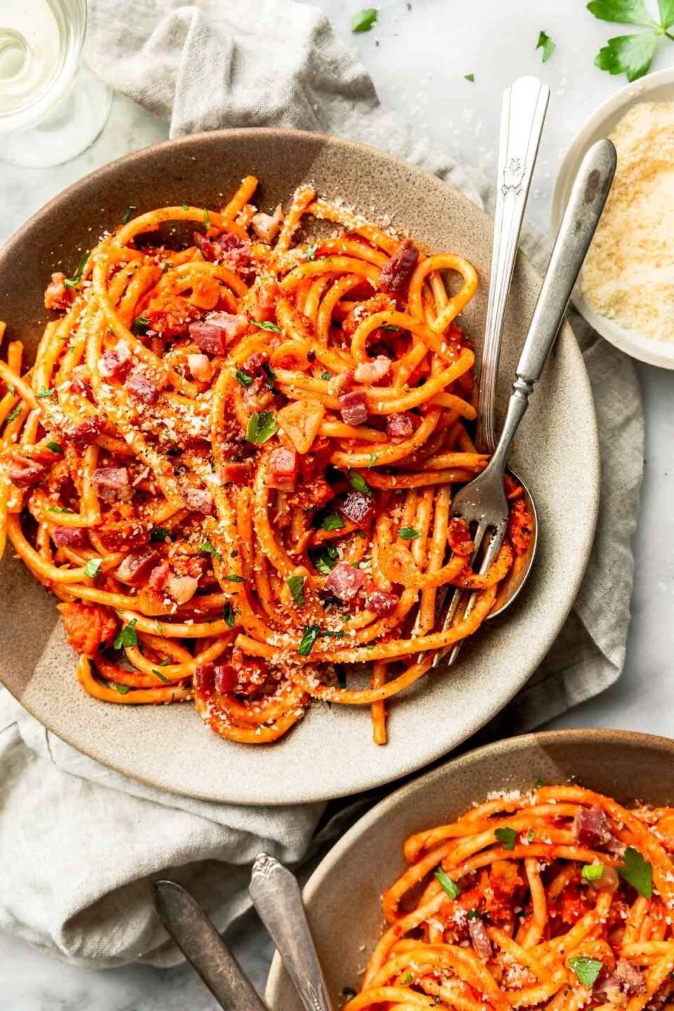 An overhead shot of a serving of bucatini all'amatriciana in a stoneware bowl on a white marbled surface. The bowl is surrounded by a second bowl of pasta, a beige cloth. glass of white wine and a dish of parmesan.