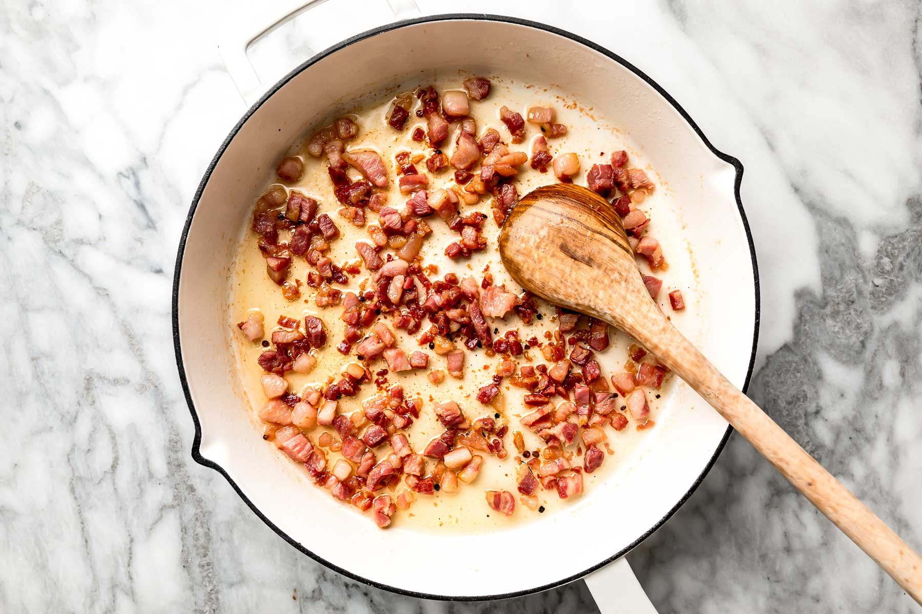 An overhead shot of guanciale rendering in a white skillet with a wooden spoon atop a white marbled surface.
