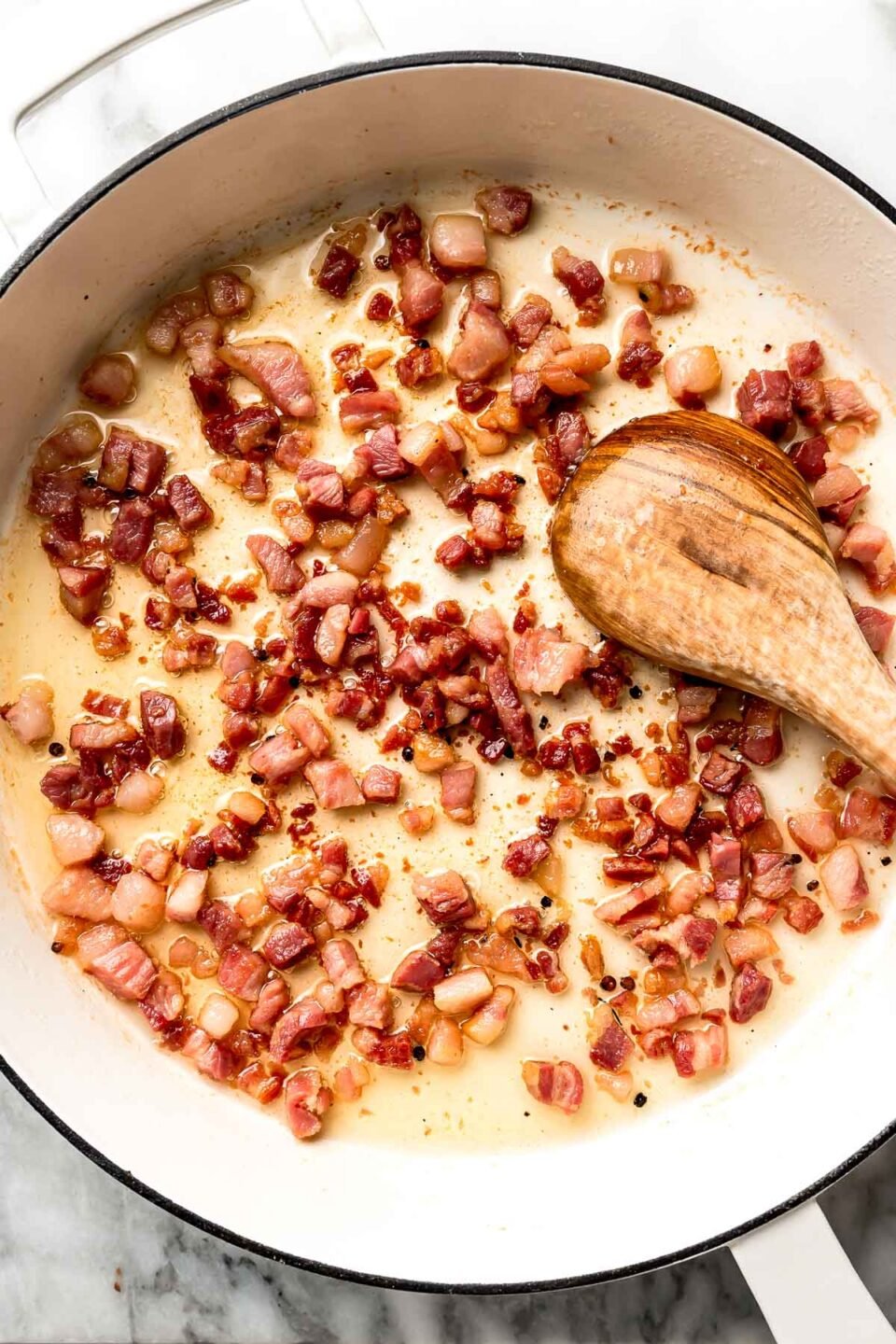 An overhead shot of guanciale rendering in a white skillet with a wooden spoon atop a white marbled surface.