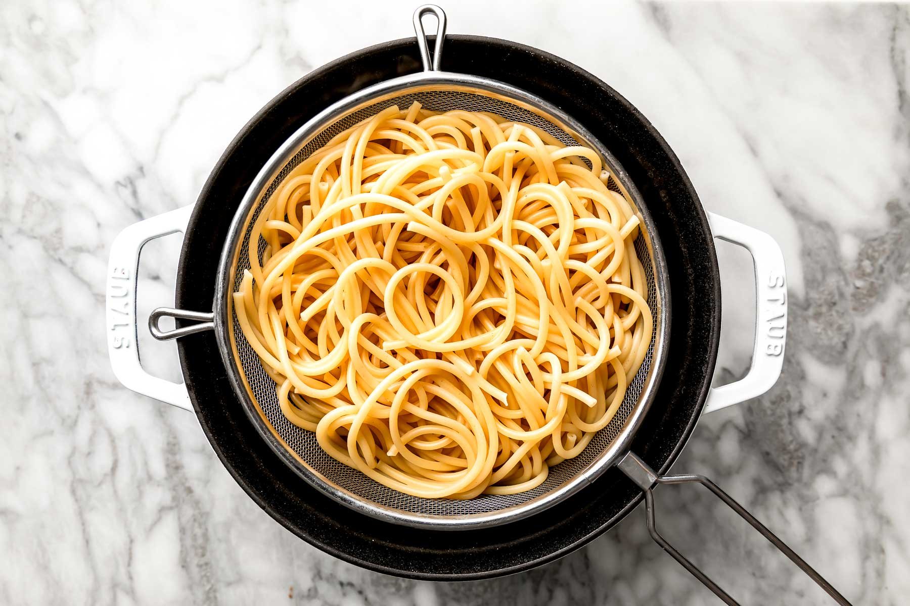 An overhead shot of cooked bucatini in a strainer over a large white pot on a white marbled surface.