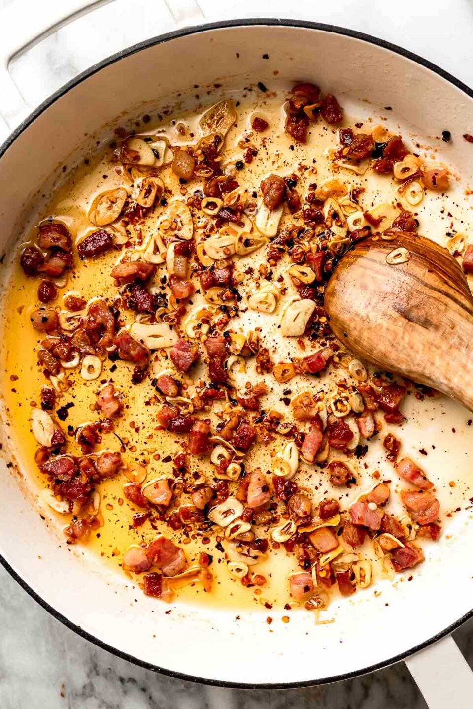 An overhead shot of pancetta and aromatics simmering in olive oil in a white skillet atop a white marbled surface.