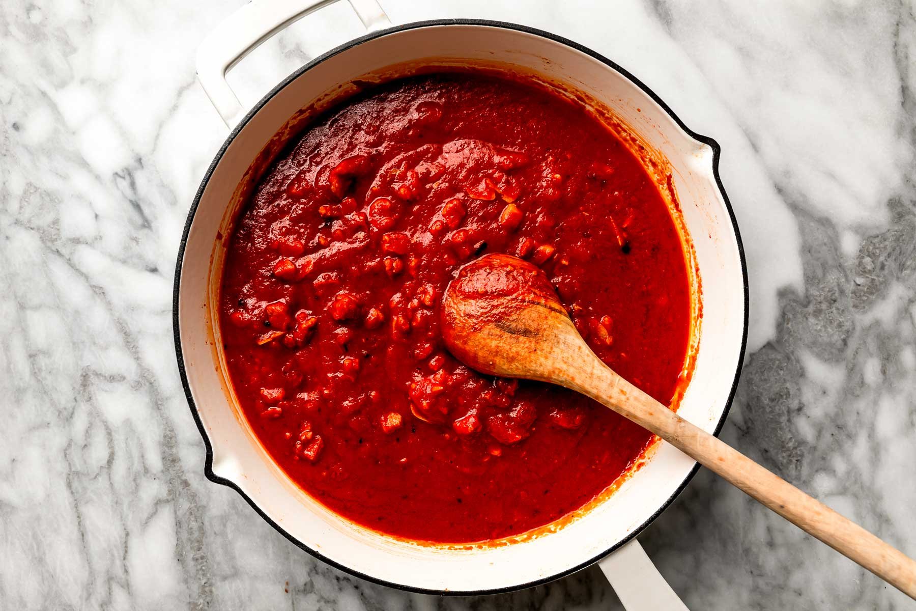An overhead shot of tomato sauce in a white skillet with a wooden spoon, sitting atop a white marbled surface.