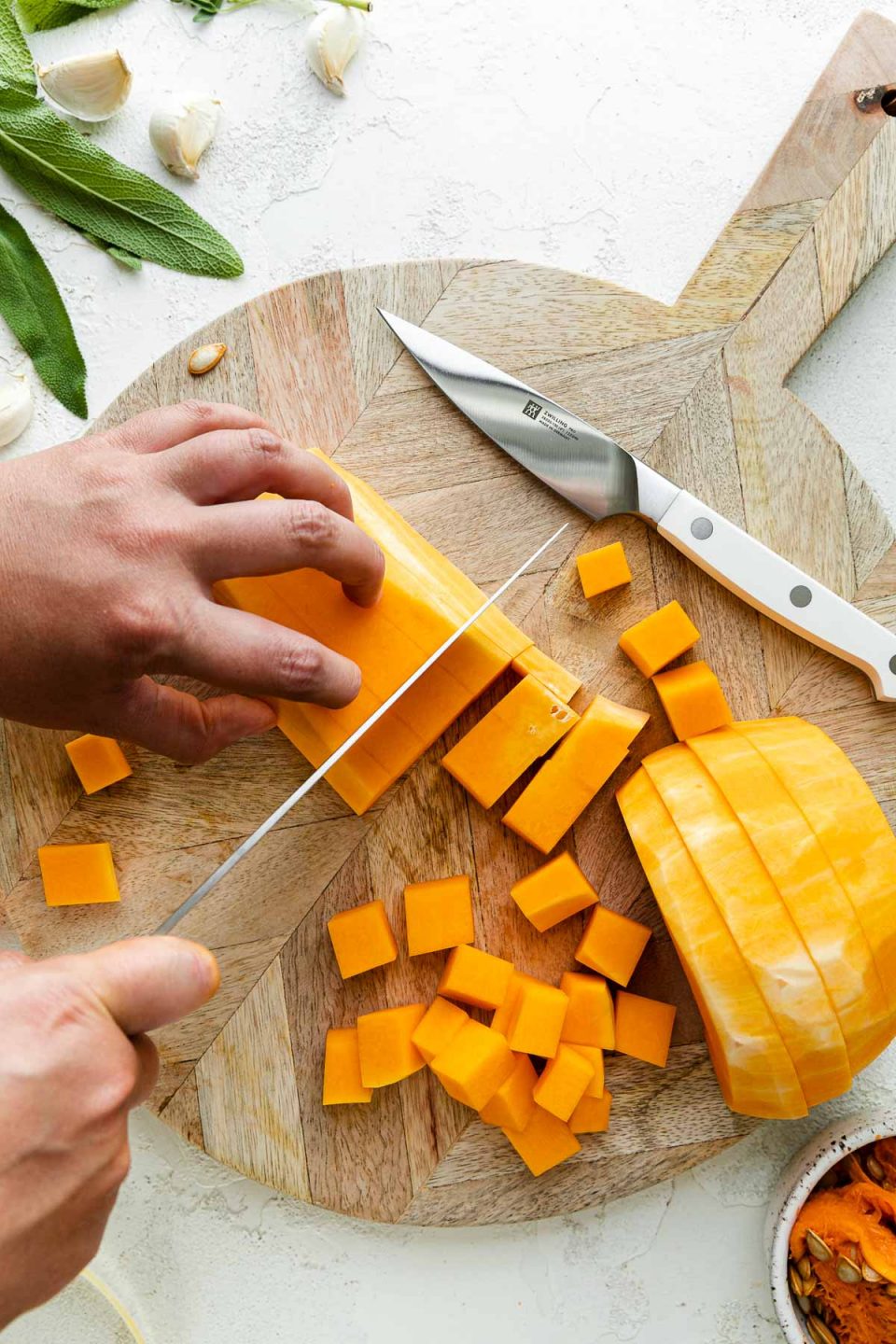 A peeled and cored butternut squash rests on top of a round wooden cutting board alongside a Zwilling Pro Le Blanc pairing knife. A woman's hands hold the top portion of a group of cut butternut squash, while using a knife to cut the squash into cubes with her other hand. Cubes of butternut squash rest on the cutting board and the cutting board sits atop a white textured surface while butternut squash peelings, fresh herbs, and cloves of garlic surround the cutting board.