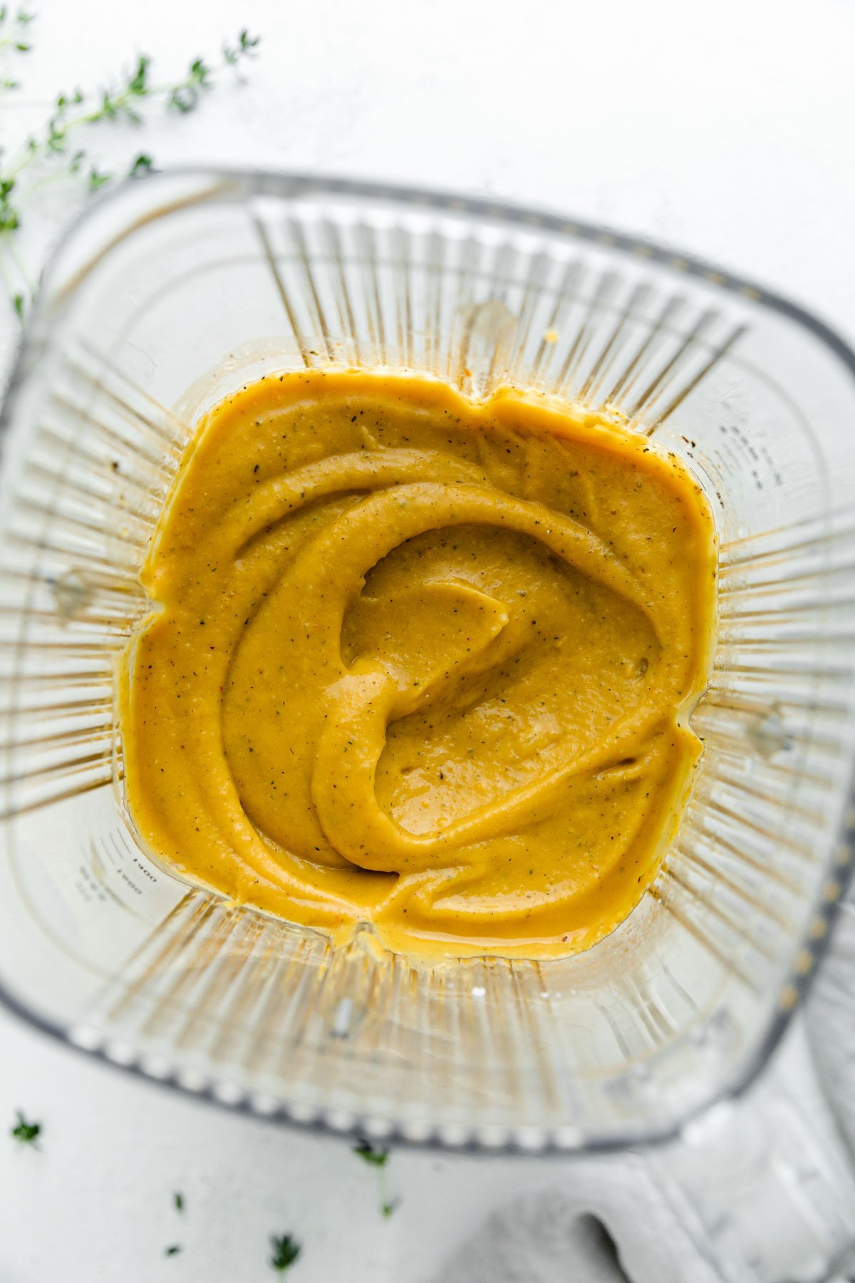 Blended creamy butternut squash pasta sauce ingredients inside of a Zwilling Enfinigy Blender. The blender carafe sits atop a white textured surface with loose herbs scattered on the surface.