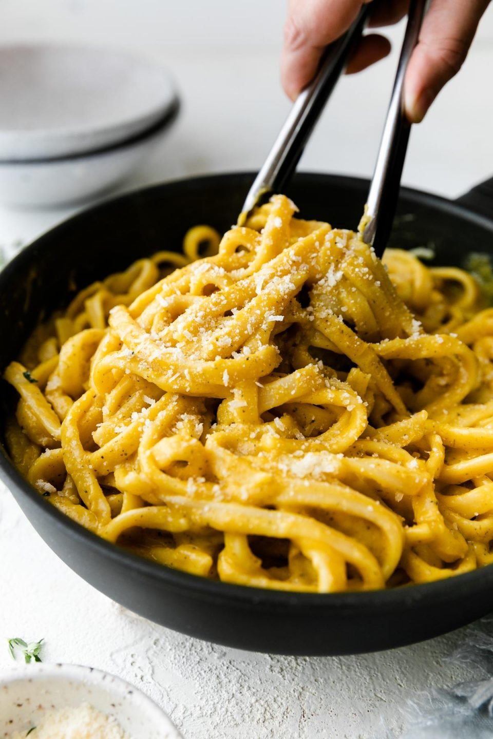 A small skillet filled with cooked linguine noodles tossed in creamy butternut squash pasta sauce. The noodles have been garnished with freshly grated parmesan and ground black pepper. A woman's hand picks up some of the pasta using a set of tongs. The skillet sits atop a white textured surface with an empty pasta bowl in the background and a small speckled bowl filled with grated parmesan cheese in the foreground.