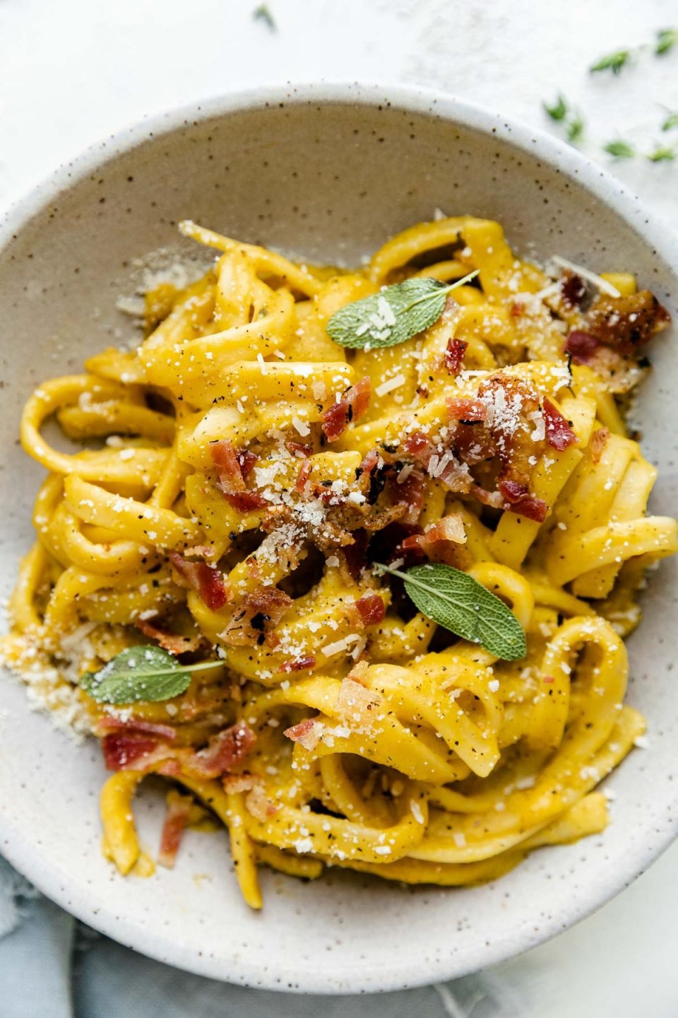 An overhead shot of creamy Butternut Squash Pasta shown in speckled ceramic pasta bowl, topped with crispy bacon, grated parmesan, ground black pepper, and fresh sage leaves. The bowl sits atop a white textured surface surrounded by a light blue linen napkin.