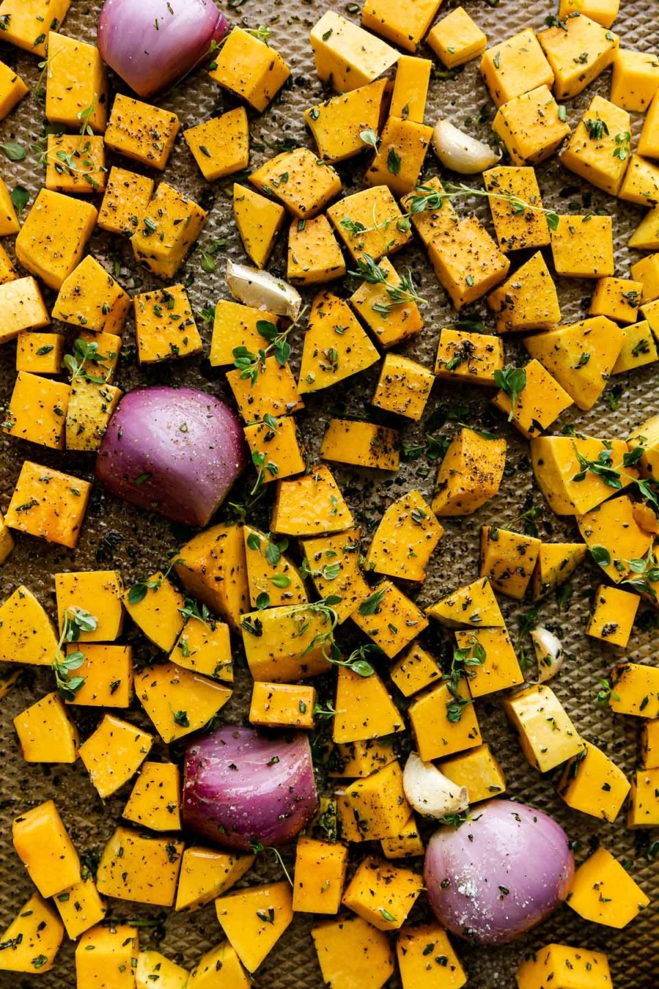 Cubed butternut squash, shallots, and garlic seasoned with olive oil and fresh herbs on a baking sheet before roasting.