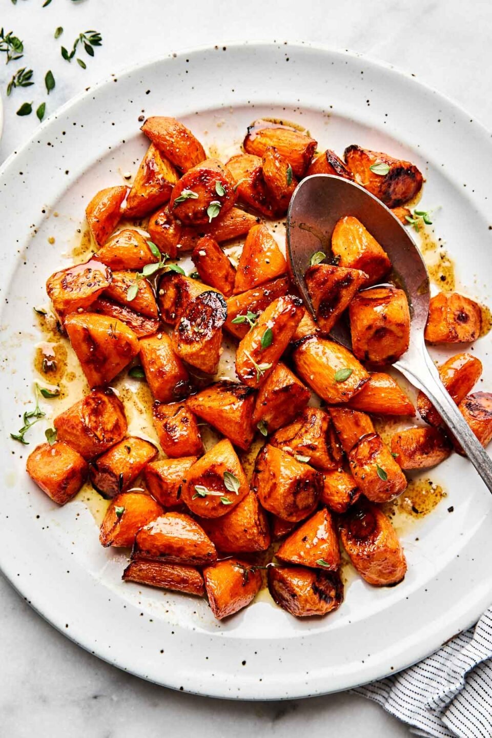 An overhead shot of roasted caramelized carrots topped with thyme leaves on a white plate atop a white marbled surface.