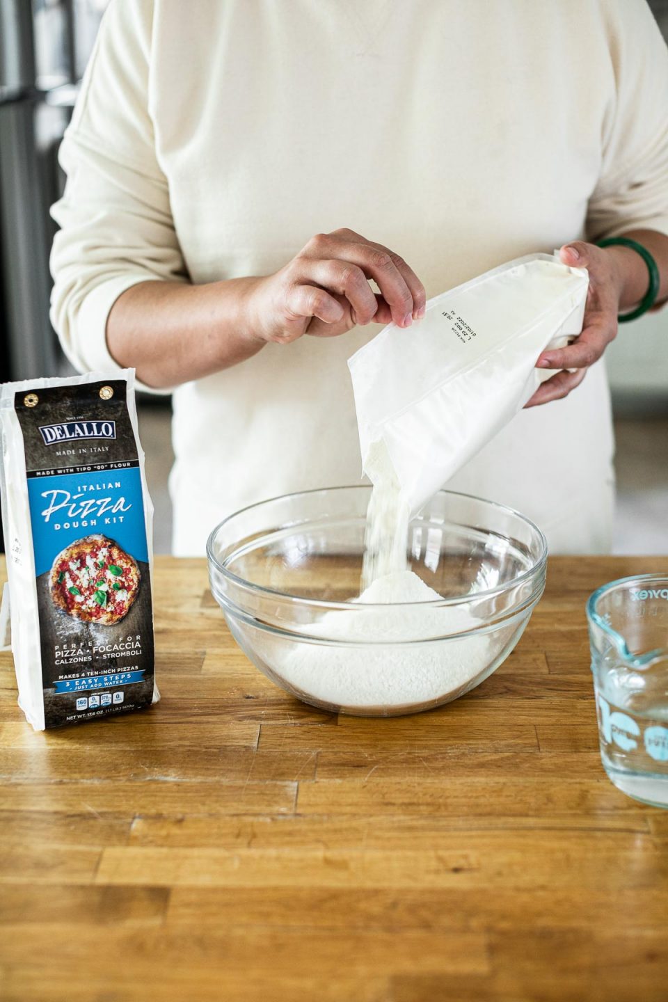 Mixing DeLallo Pizza Dough Kit: Jess, shown in a white sweatshirt, stands behind a butcher block counter, pouring DeLallo Pizza Dough Kit into a large glass mixing bowl. Sitting next to the bowl on the counter are a wooden spoon & a package of DeLallo Pizza Dough Kit.