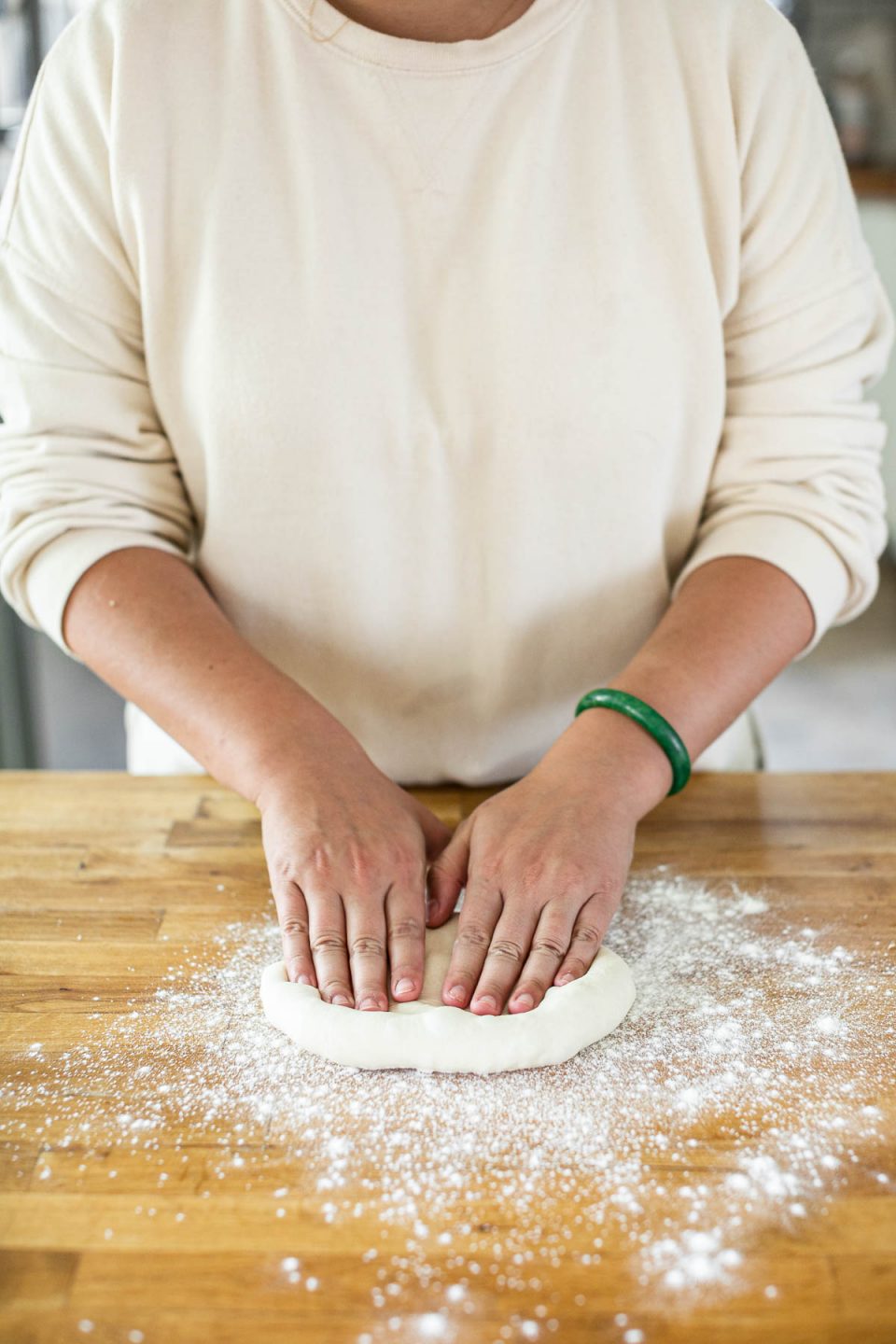 How to form pizza crust: Jess, shown in a white sweatshirt, stands behind a butcher block counter dusted in flour. Jess' fingers are pressed into a pizza dough ball, forming it into crust.