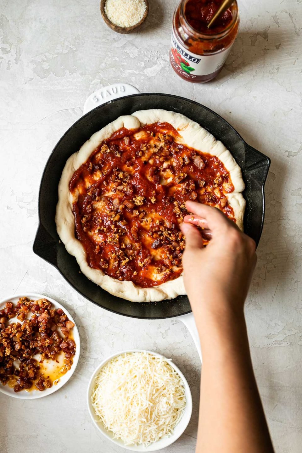 A woman's hand reaching into the frame with a gold spoon, spreading DeLallo pizza sauce over pizza crust formed in a white Staub cast iron skillet. The skillet sits on a white surface, surrounded by parmesan cheese, a jar of DeLallo pizza sauce, fried pancetta & garlic, & shredded Italian cheese.