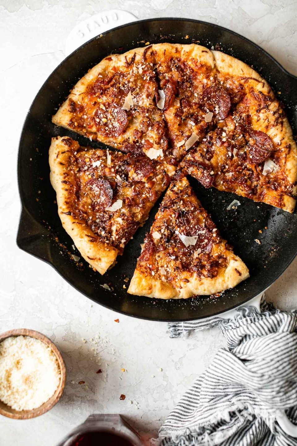 Baked Pizza Amatriciana in white Staub cast iron skillet. The pizza is cut into 6 slices. The skillet sits on a white surface, surrounded by a striped linen napkin, a glass of red wine & a small wooden bowl of grated parmesan cheese.