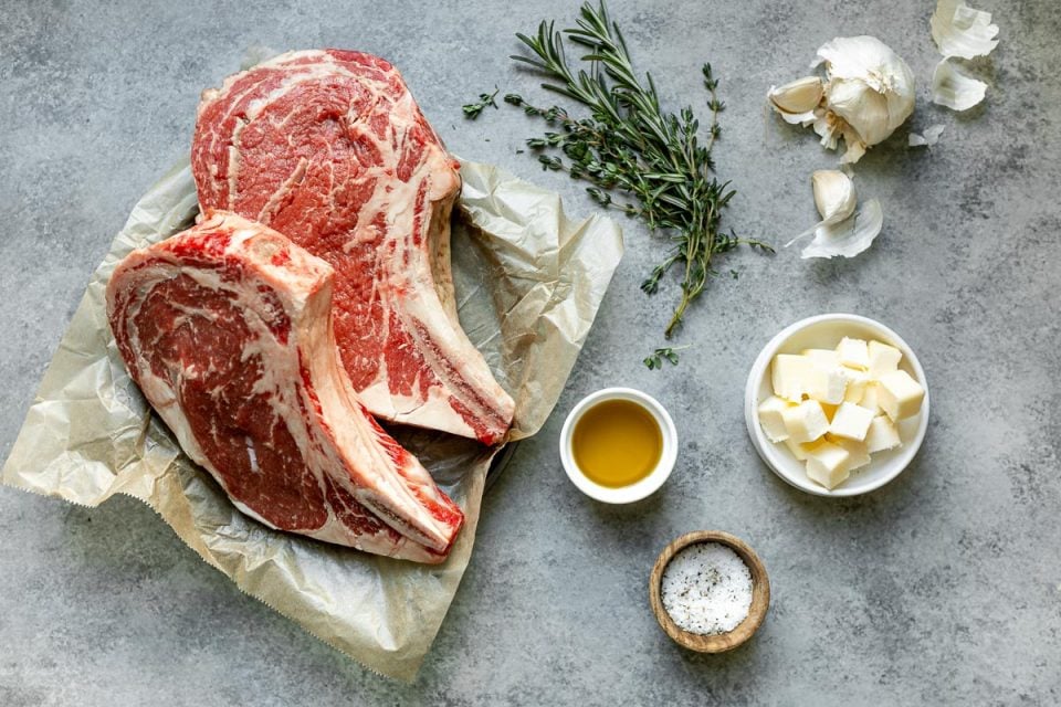 An overhead shot of ingredients arranged on a light gray surface: bone-in ribeye steaks, avocado oil, salt and pepper, cubed butter, garlic, and fresh herbs.