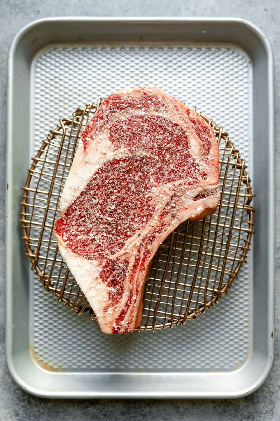 An overhead shot of a bone-in ribeye, seasoned generously with salt, placed atop a wire rack on a small baking sheet.