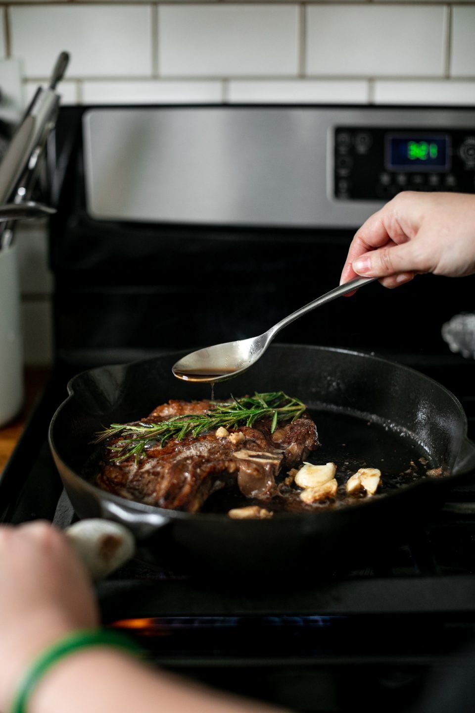 A woman's hands hold handle of skillet and spoon, spooning browned butter over the steak, which is topped with fresh herbs and crushed garlic cloves.