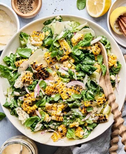 An overhead shot of charred sweet corn Caesar salad in a large shallow white bowl with a wooden serving spoon alongside a striped cloth on a light blue surface. The bowl is also surrounded by halved and juiced lemons, a jar of dressing, and bowls of black pepper and parmesan cheese.