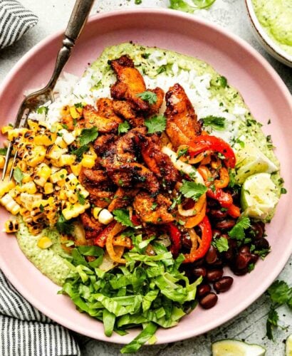 An overhead shot of an assembled fajita bowl on a light pink plate atop a striped cloth on a light grey surface: avocado crema, fajitas, lettuce, corn, pico de gallo, rice and black beans, topped with cilantro.