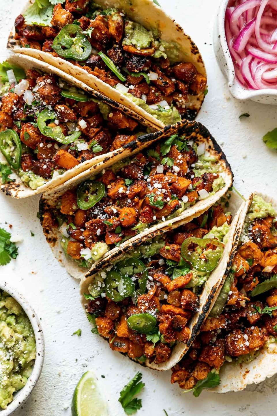 Overhead view of chicken al pastor tacos, pressed together side-by-side on a white plaster surface. The tacos are surrounded by fresh cilantro leaves, lime wedges, & mashed avocado & pickled red onions in separate white bowls.