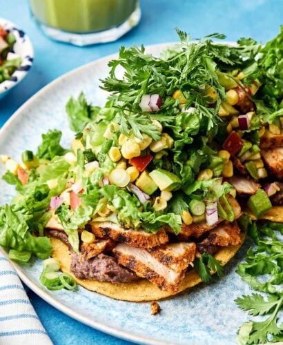 An overhead shot of two tostada shells topped with grilled chicken and tostada salad on a blue speckled plate atop a blue surface. A jar of cilantro lime vinaigrette sits in the background.