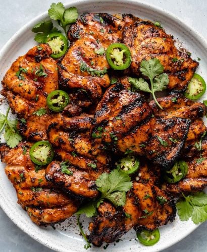 Grilled chili lime chicken thighs shown on a white speckled plate atop a light blue surface. The chicken is garnished with fresh cilantro leaves, sliced jalapeño, & ground black pepper.