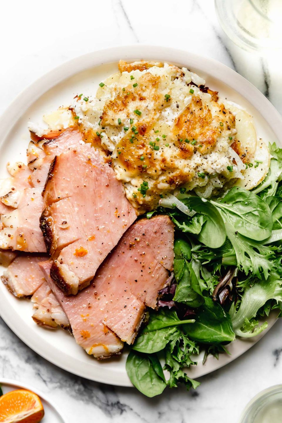 Thick slices mapxle glazed baked ham, plated on a dinner plate with greens & au gratin potatoes.