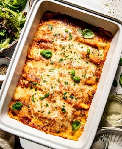 An overhead shot of crepe manicotti in a large casserole dish garnished with fresh basil. The dish sits on a white textured surface alongside dishes of parmesan and pepper, a plate with a fork and spoon, and a side salad.
