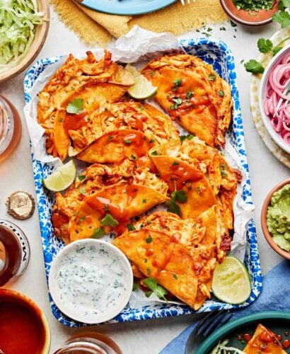 An overhead shot of baked buffalo chicken tacos garnished with limes, cilantro and buffalo sauce alongside a bowl of ranch on a blue and white patterned platter. The platter is surrounded by shredded lettuce, red onions, guacamole, cilantro, buffalo sauce and plates of tacos on a white surface.