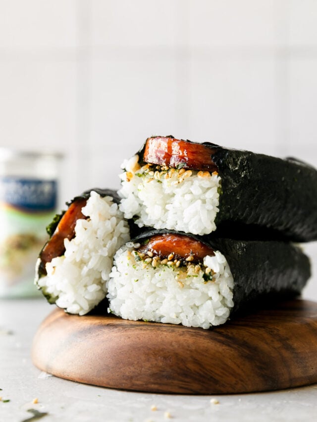 Three Hawaiian spam musubi are arranged atop a wooden serving platter. The platter sits atop a creamy white textured surface and a container of Furikake seasoning sits out of focus in the background.