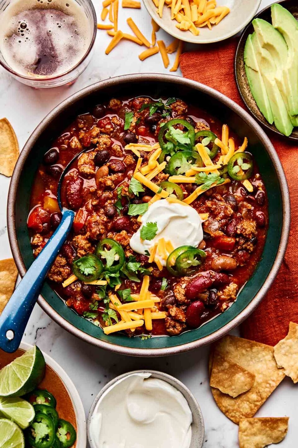 An overhead shot of turkey chili in a dark green bowl atop a white surface. The bowl is surrounded by an orange cloth, tortilla chips, a bottle of beer and plates of sliced avocado and lime wedges.