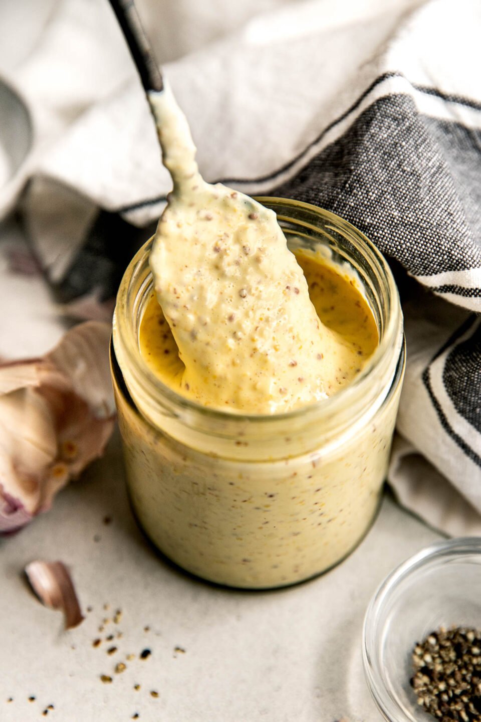 An angled shot of a spoon lifting sauce from a glass jar of dijonnaise on a white textured surface alongside garlic, a dish of pepper and a dish towel.