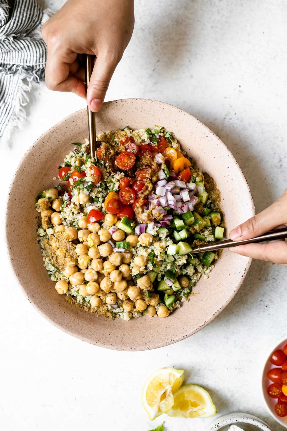 Falafel-ish Quinoa Salad in a large, pink serving bowl. A woman's hands hold the bowl up off of a white surface. Surrounding the salad are a linen napkin, & some lemon wedges.
