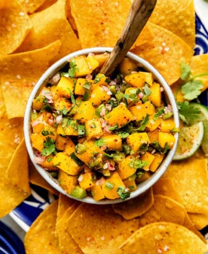 A close-up overhead shot of a small bowl of mango salsa surrounded by tortilla chips on a blue and white platter.