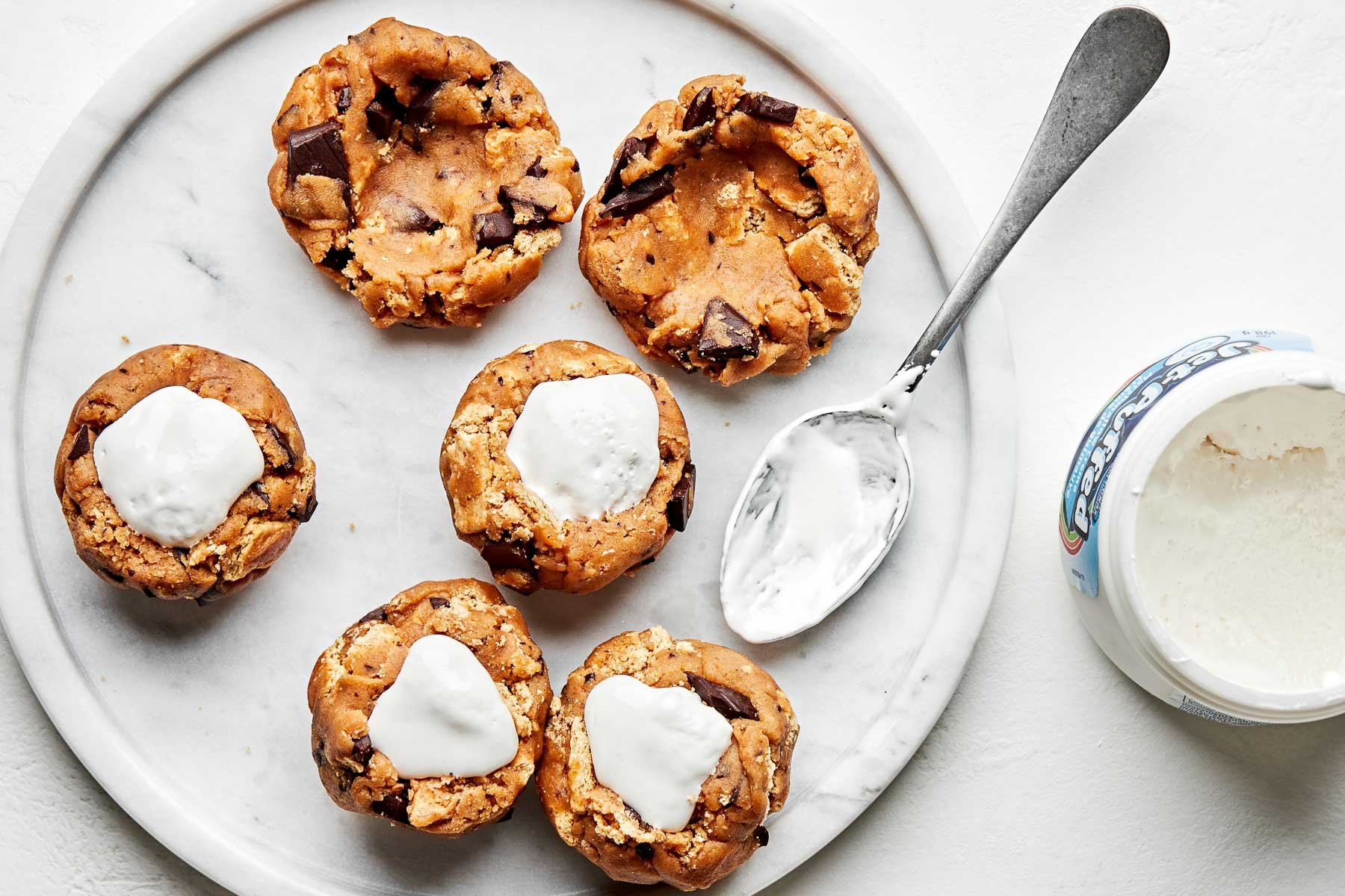 An overhead shot of several rounds of cookie dough that have had marshmallow fluff spooned into the center. Two plain cookie dough rounds sit alongside them, as well as a spoon and an open jar of marshmallow fluff.