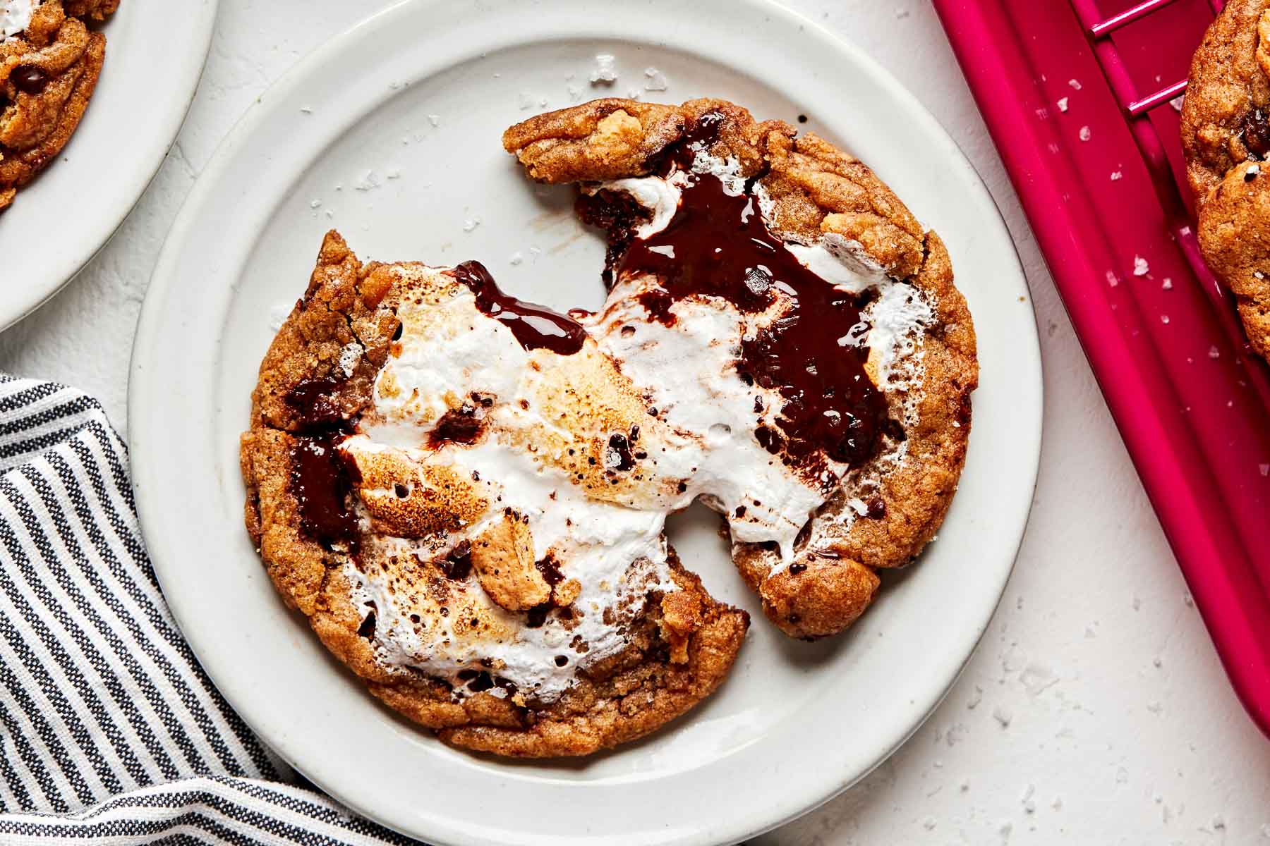 An overhead shot of a s'mores cookie that has been broken partially in half, showing the gooey marshmallow fluff in the center. The cookie sits on a white plate on a white surface, alongside a striped dish towel and a pink baking sheet.
