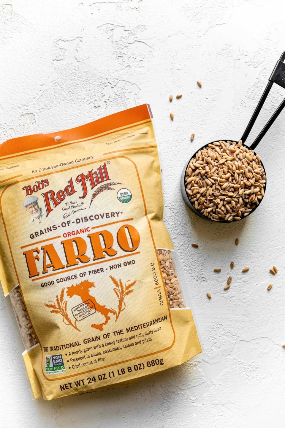 Bag of Bob's Red Mill farro on a white surface next to black measuring cup with farro in it.