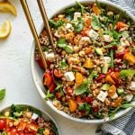 Greek farro salad in ceramic serving dish atop a white surface. The farro salad is tossed & topped with fresh basil leaves & has serving spoons nestled in it. The bowl is surrounded by a striped gray linen napkin, a small plate of Greek farro salad & some lemon wedges & fresh herbs.