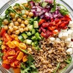Greek farro salad in large clear glass bowl atop a white surface. The farro salad ingredients are grouped in the bowl - lemony red onions, chickpeas, cucumber, tomatoes, farro, feta, etc.