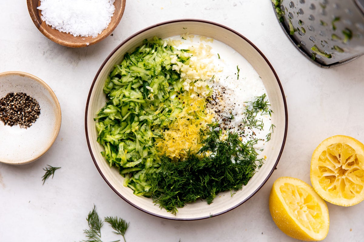 How to make greek pasta salad, step 3: Prep the creamy tzatziki dressing. Plain Greek yogurt, lemon zest & juice, garlic, fresh dill, olive oil, kosher salt, and ground black pepper are added to a cream colored bowl that sits atop a creamy white textured surface. A small wooden pinch bowl filled with kosher salt, a small ceramic pinch bowl filled with ground black pepper, a metal box grater, loose fresh dill, and two spent lemon halves surround the bowl at center.