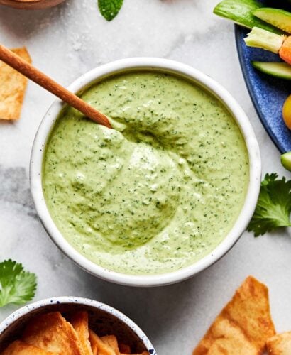 A close-up overhead shot of green tahini sauce in a white bowl atop a white marbled surface. A small bowl of pita chips, a bowl of salt, several sprigs of cilantro and a blue plate of cherry tomatoes, cucumbers and carrots sit alongside it.