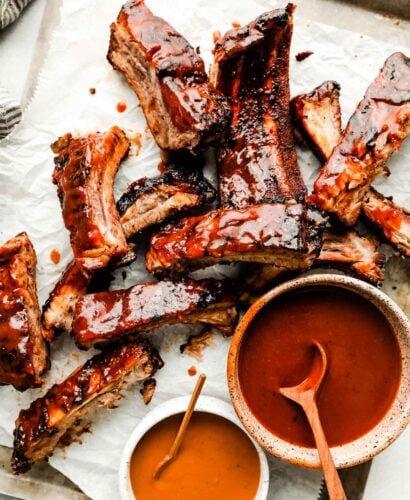 An overhead shot of individual grilled ribs on a paper-lined sheet pan atop a white surface. Two bowls of BBQ sauce sit alongside them.