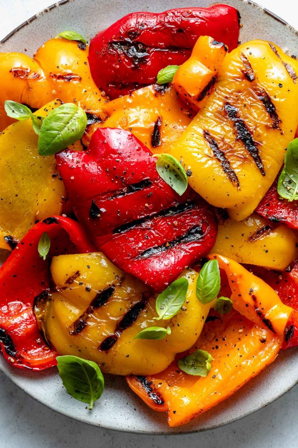 Close up of yellow, orange, and red grilled bell peppers with char marks arranged on a ceramic plate. The grilled bell peppers are garnished with kosher salt, ground black pepper, and fresh herbs. The platter sits on top of a white and gray marble surface.