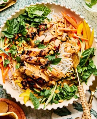 A bowl of grilled chicken strips over rice noodles, topped with peanut sauce, chopped peanuts, fresh basil, mint, sliced carrots, cucumber, and yellow bell peppers, with a gold fork and knife beside the bowl.