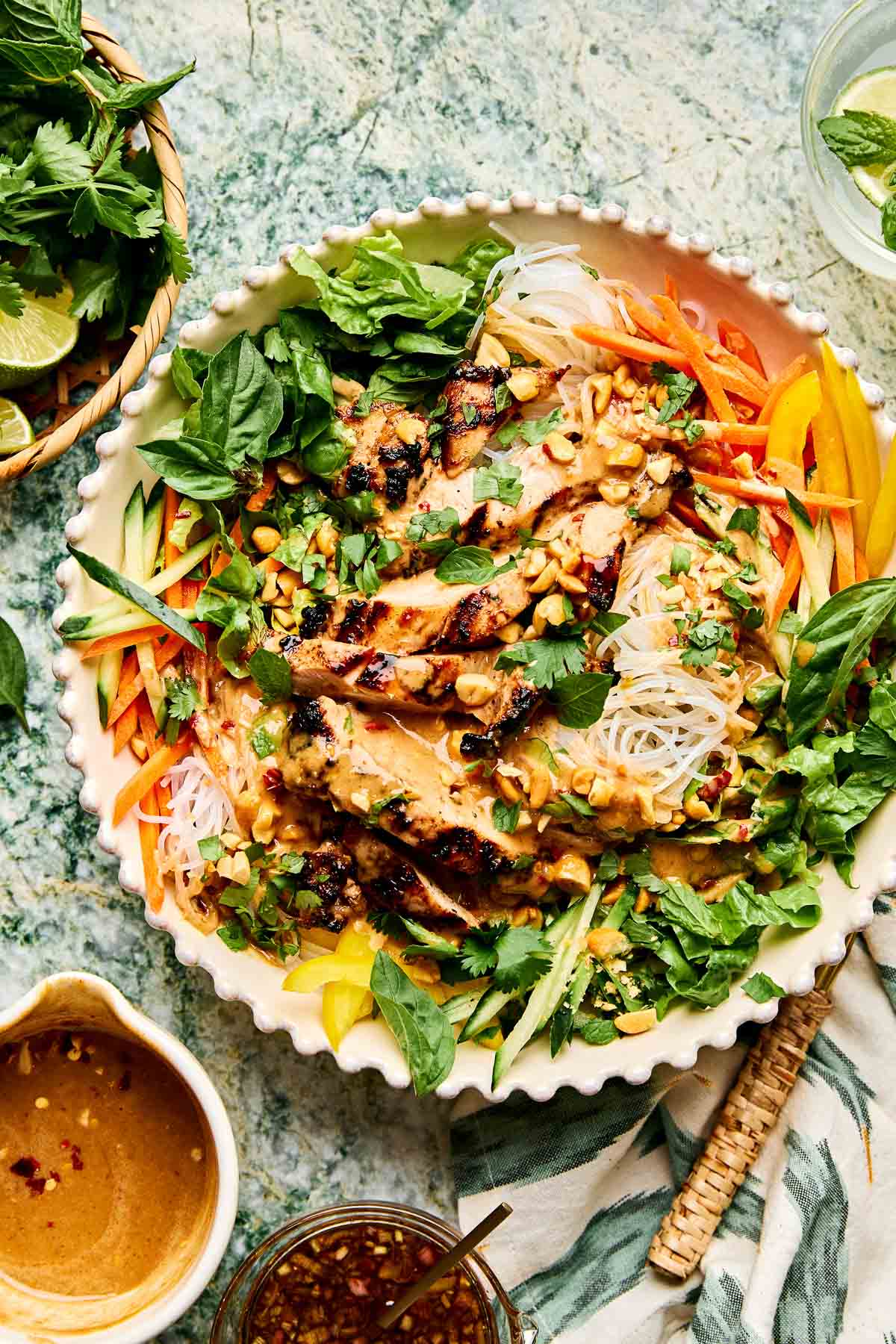 A large bowl filled with sliced grilled chicken, rice noodles, shredded carrots, bell peppers, cucumber, fresh herbs, and peanuts, all drizzled with a creamy peanut sauce, set on a green marble surface.